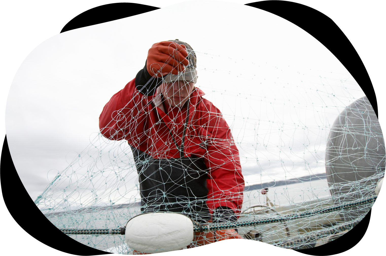 Ung tenåringsgutt som jobber på fiskebåt. Han holder i et fiskegarn. Han har på seg rød kjeledress, caps og hansker. I bakgrunnen ser vi sjøen, og det er grått vær. Foto: moodboard/Adobe Stock.
