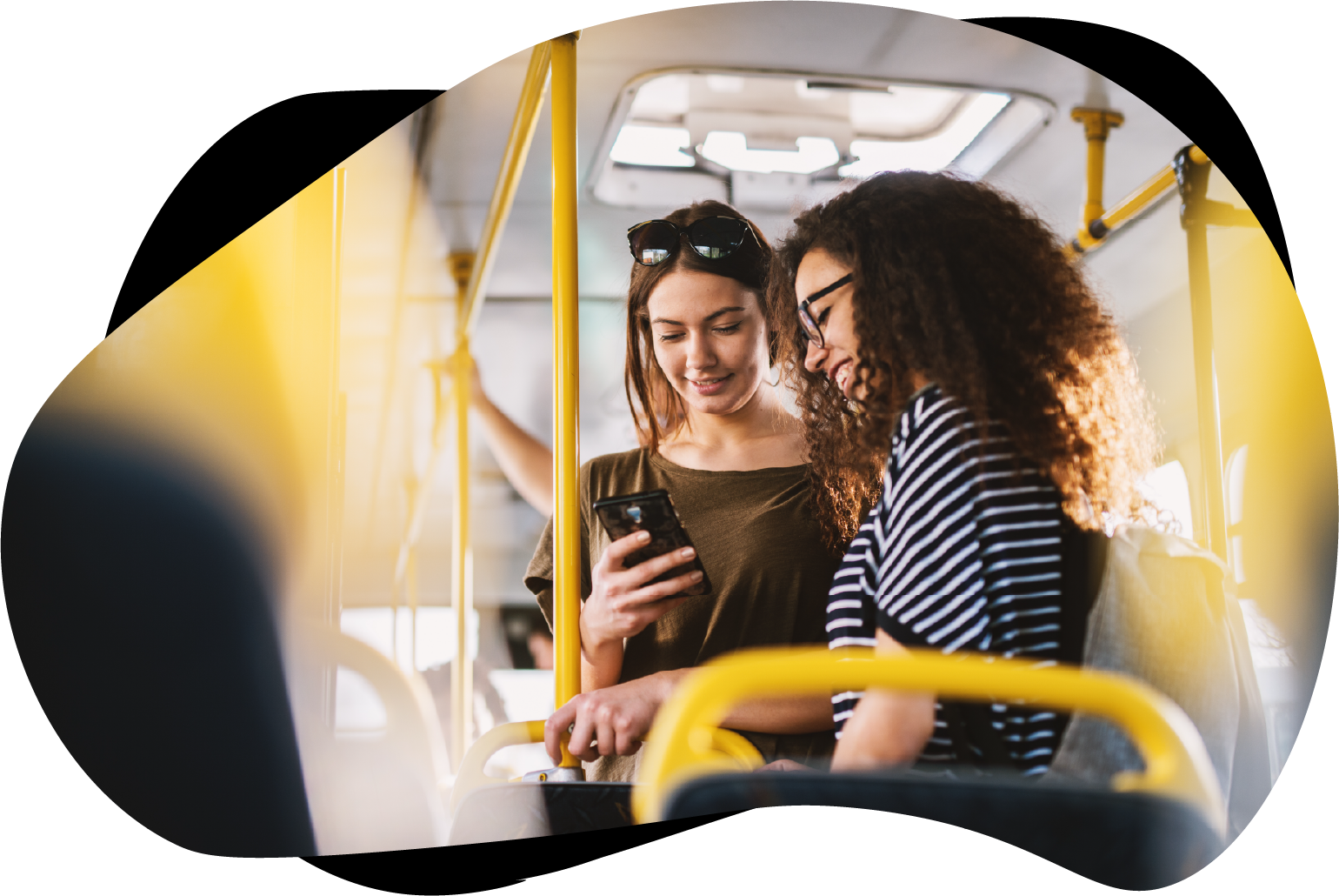 To unge jenter (tenåringer) står i midtgangen i en buss, mens de ser på telefonen og smiler. Foto: Dusan Petkovic/Adobe Stock.